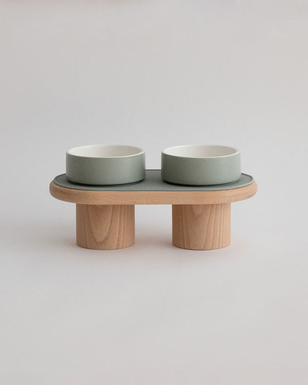 Soft sage green ceramic bowls with silicone mat on a solid wood stand with natural grain.