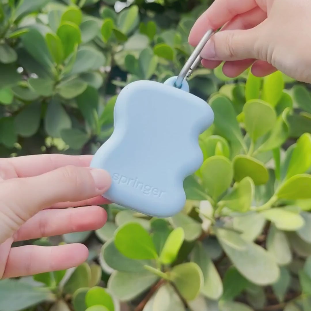 Close-up of a skyblue silicone treat dispenser clipped to a carabiner in front of green leaves, lightweight portable dog training reward dispenser.