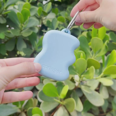 Close-up of a skyblue silicone treat dispenser clipped to a carabiner in front of green leaves, lightweight portable dog training reward dispenser.