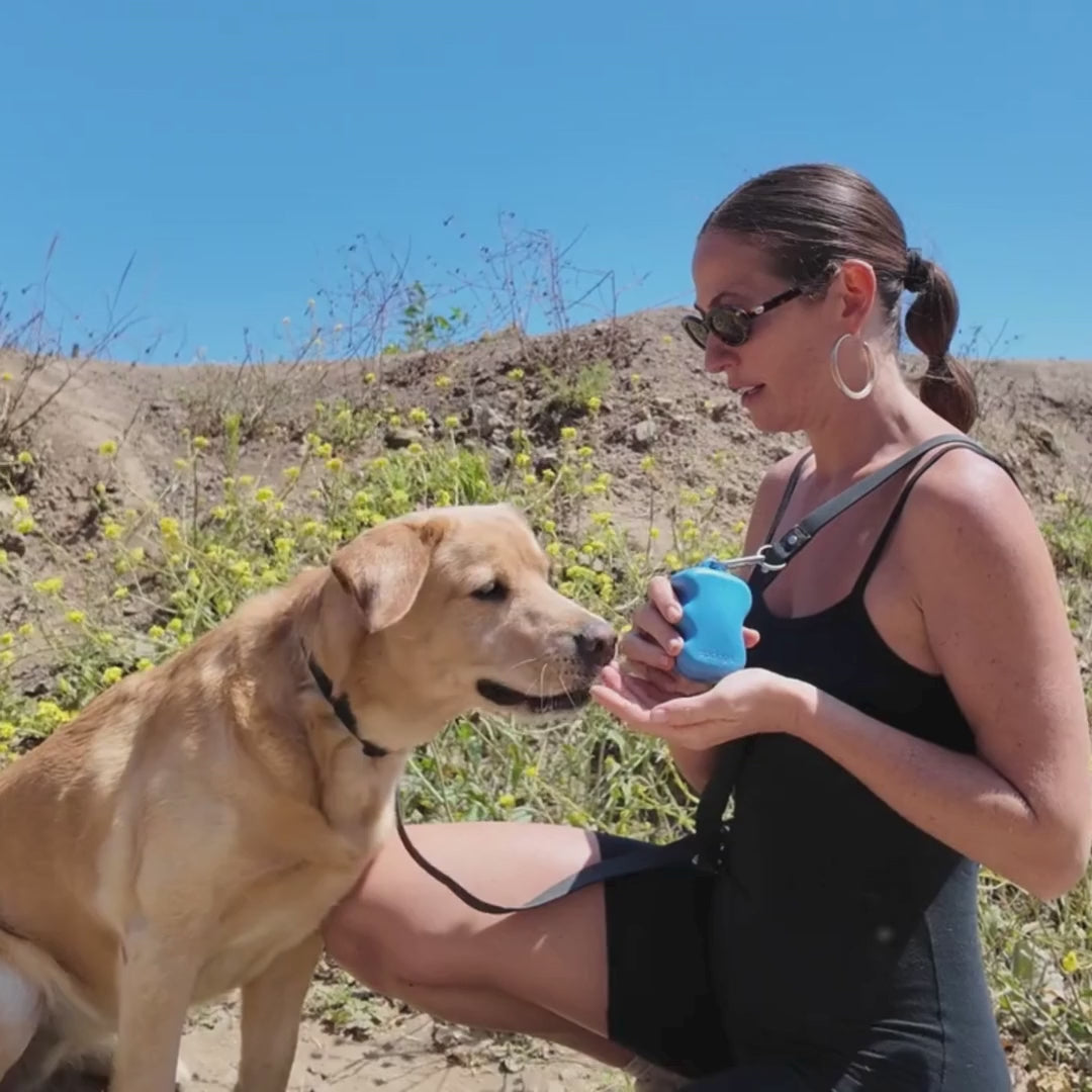 Woman giving a dog treats during outdoor training while holding a blue silicone treat dispenser clipped to a leash or bag.