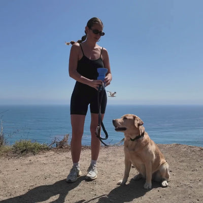 Person hiking with a dog carrying a blue travel dog water bottle, convenient hydration for outdoor walks and adventures.